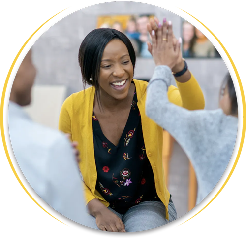A person in a yellow cardigan high-fives another individual, both appearing joyful, in an office or classroom setting.