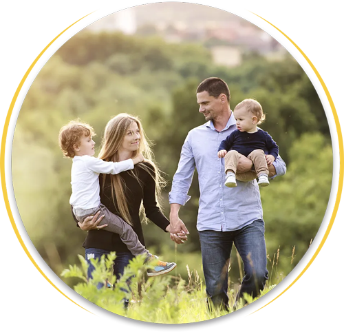 A family walks through a sunny, grassy field; a parent carries a child in each arm while holding hands.