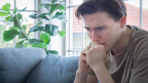 A person in a brown shirt sits on a sofa, looking downward with a worried expression, hands clasped near their chin.