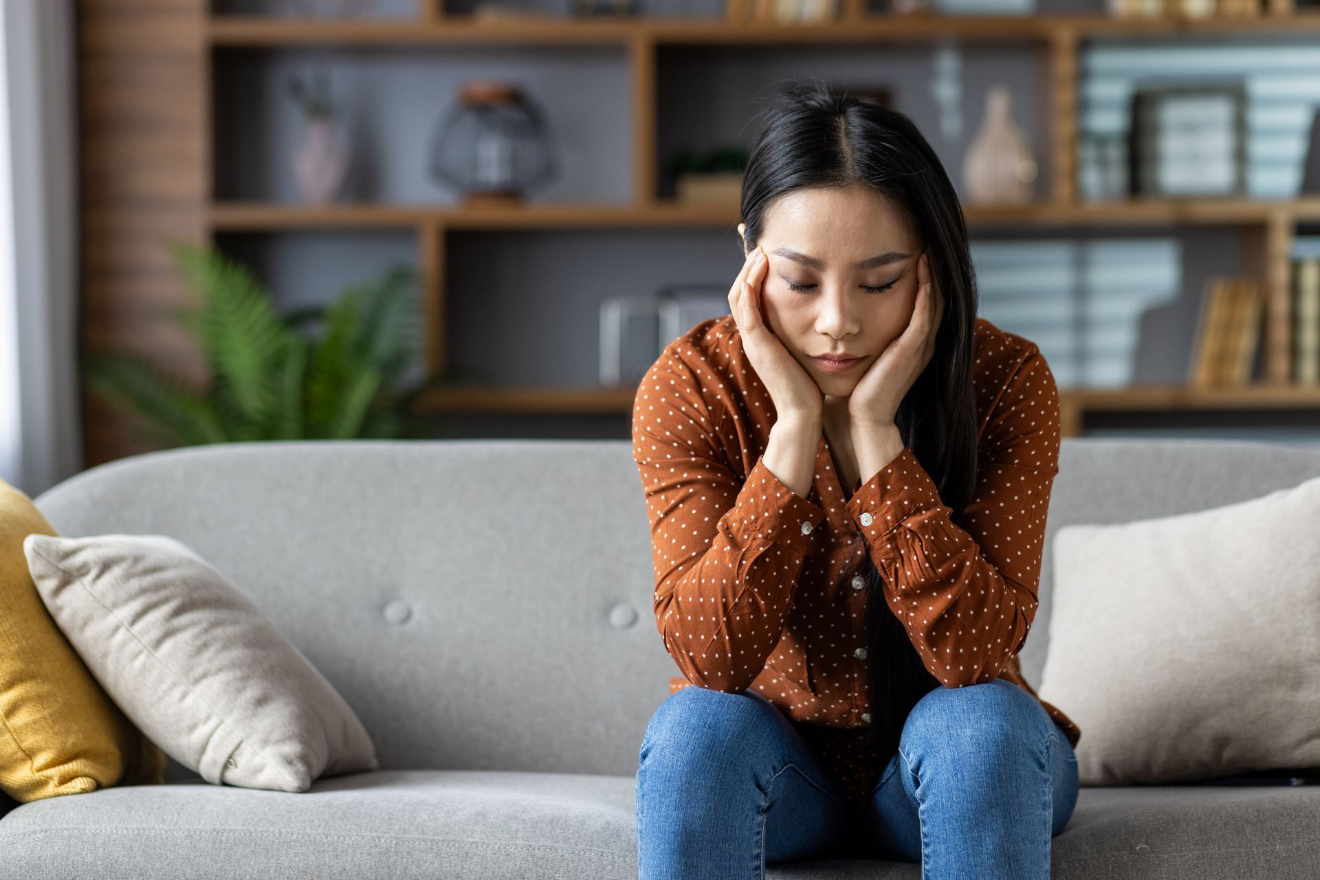 A person sits on a sofa, leaning their head on their hands with a sad, pensive expression.