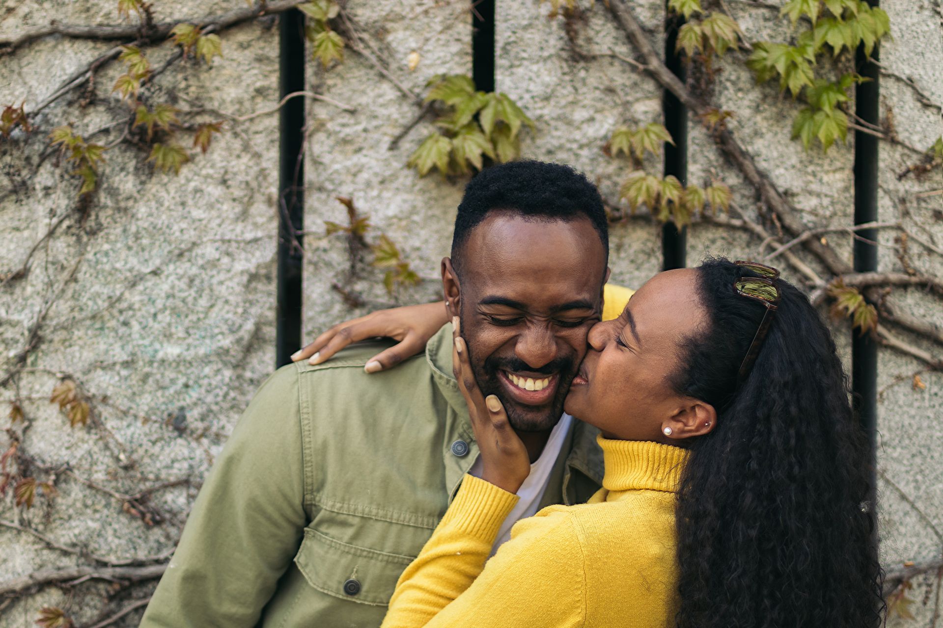 A person in a yellow sweater kisses another person on the cheek against a stone wall covered in vines.
