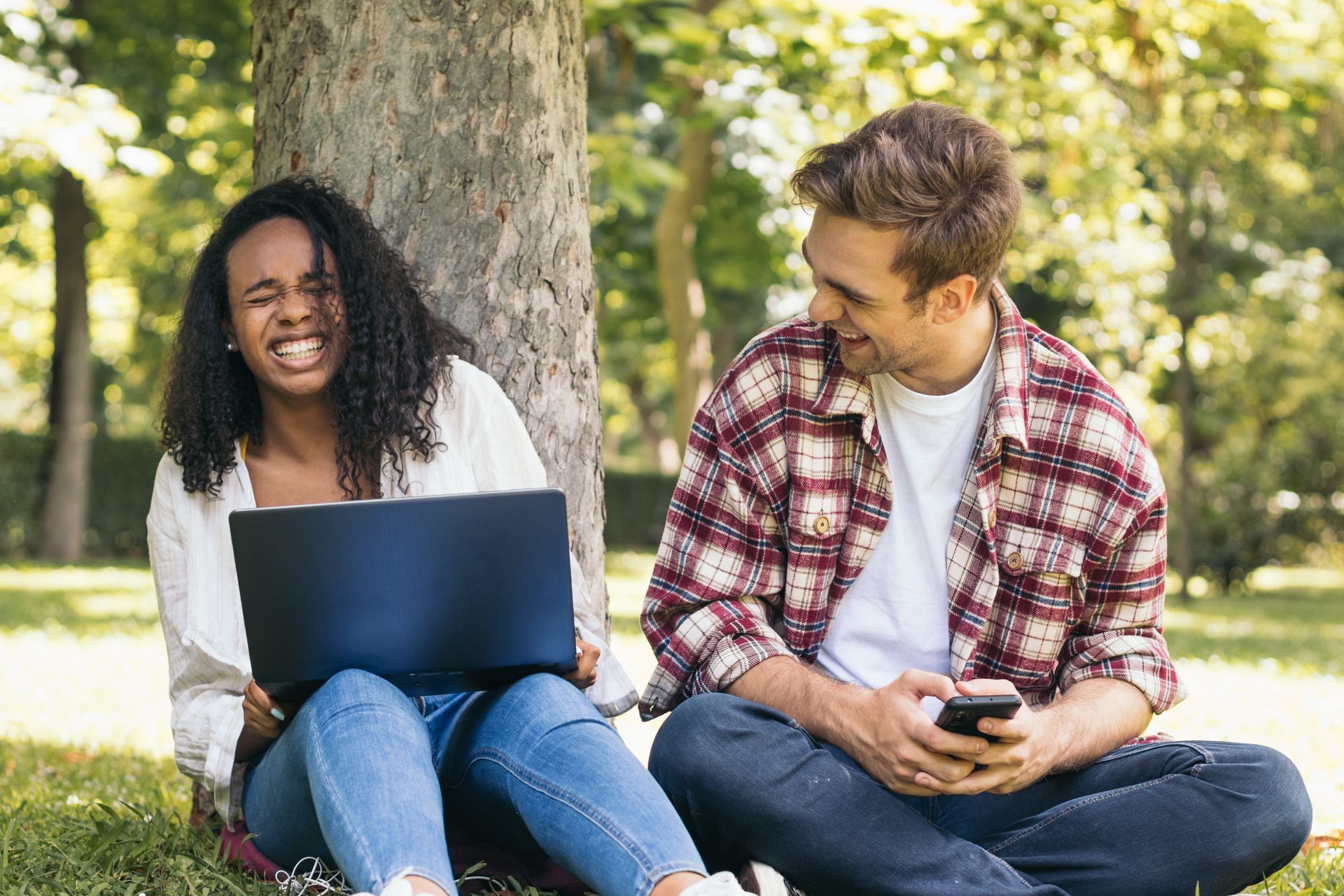 Two students sit under a tree in a park, laughing together while using a laptop and a smartphone.