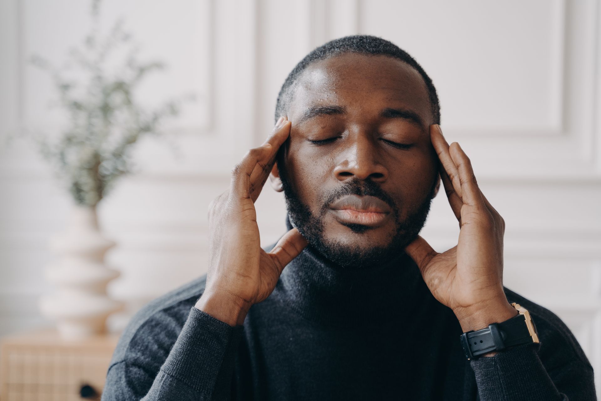 A person in a dark sweater holding their temples with eyes closed in a calm, indoor setting.