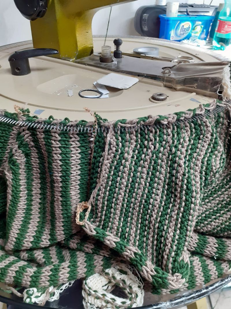Tessuto lavorato a maglia a righe verdi e beige in lavorazione, su un tavolo rotondo con una macchina da cucire e i relativi materiali.