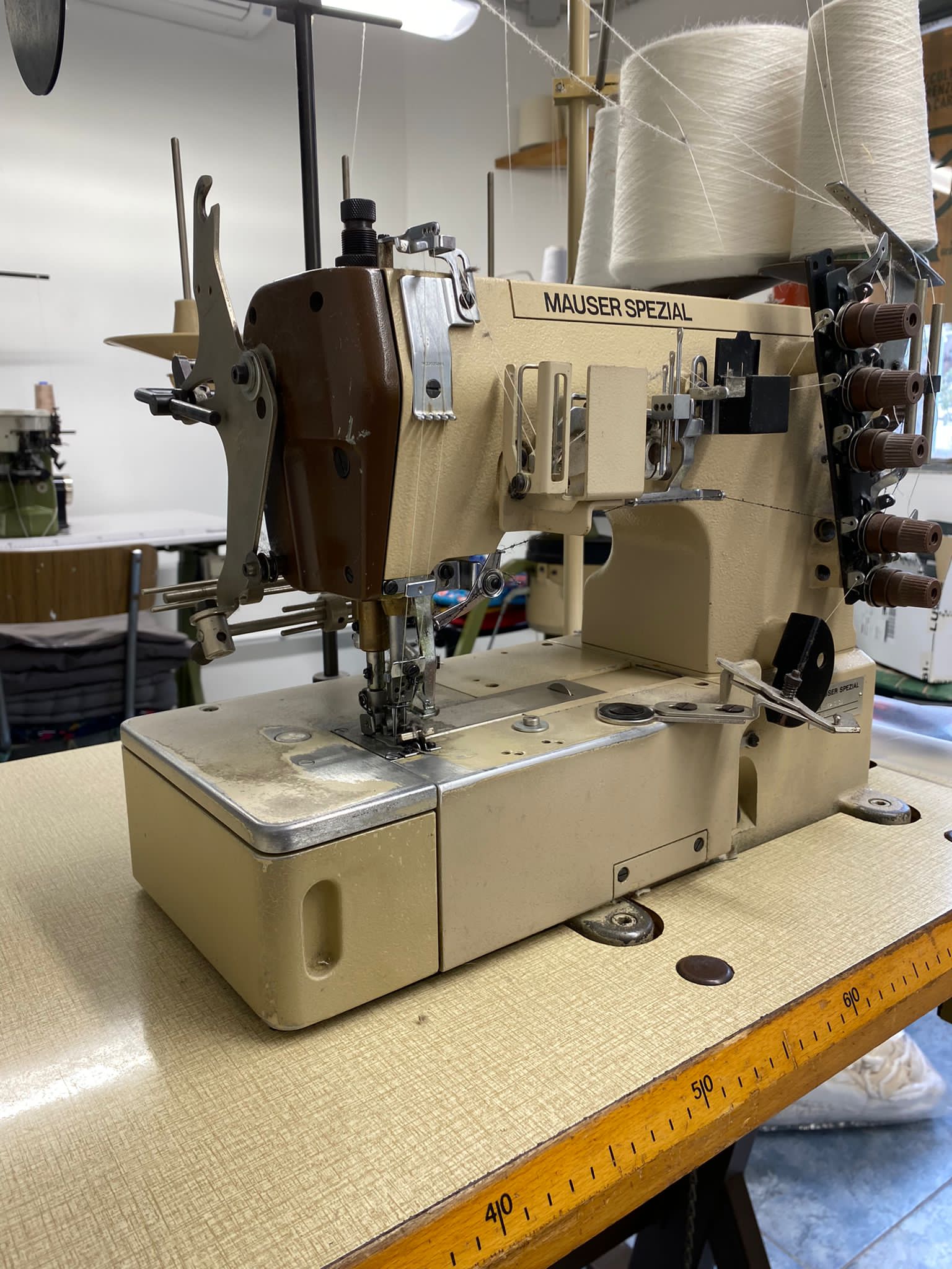 Macchina da cucire industriale beige su un tavolo in un'officina, con rocchetti di filo sullo sfondo.