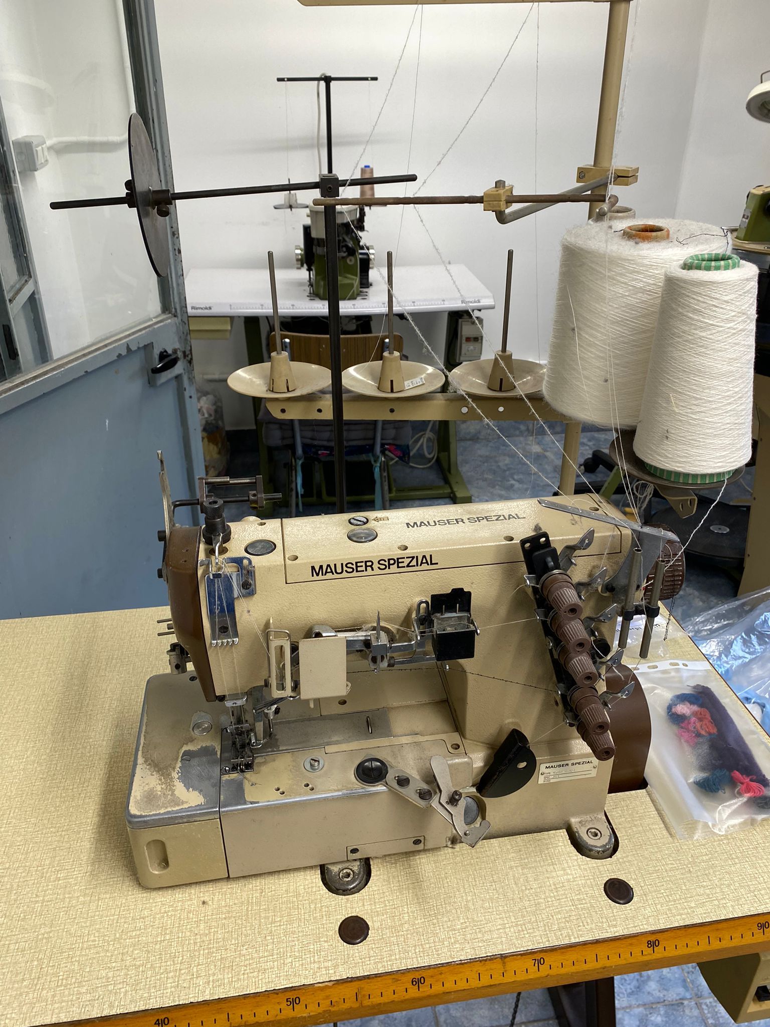 Macchina da cucire industriale su un tavolo, con rocchetti di filo e altre attrezzature in un ambiente da officina.