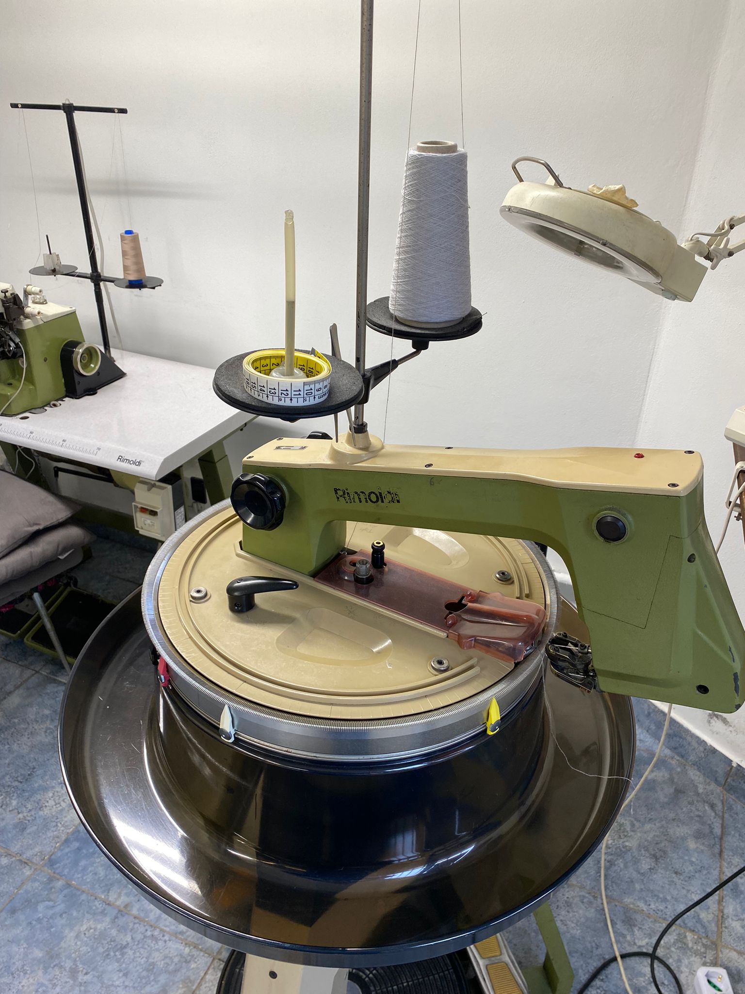 Macchina circolare per maglieria con rocchetto di filo, lampada da lavoro e tavolo da lavoro sullo sfondo.