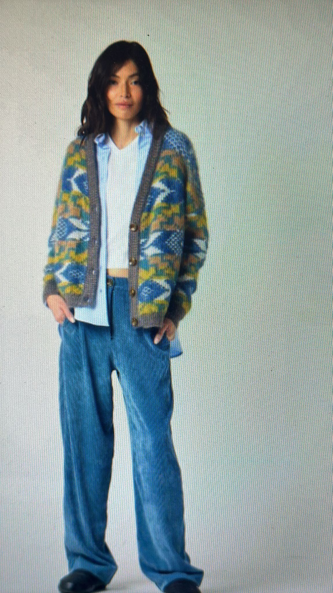 Donna che indossa un cardigan fantasia, pantaloni scozzesi blu e una camicia con colletto, con le mani in tasca.