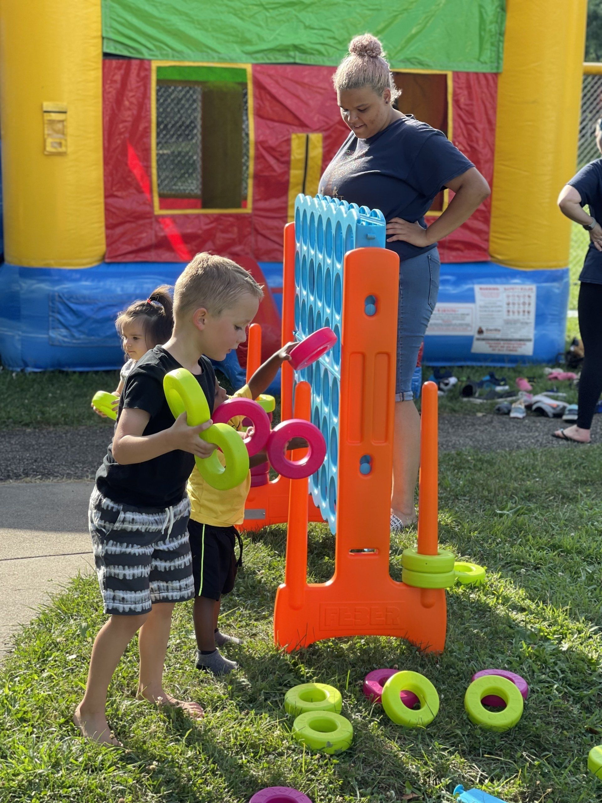three kids playing a large connect four game at a preschool event next to a bouncy house