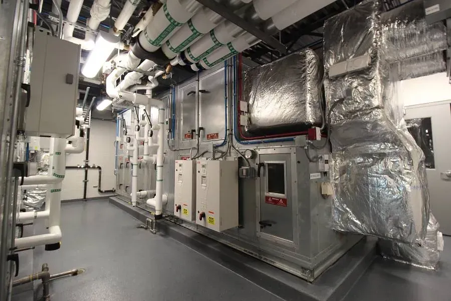 Industrial HVAC/mechanical room with large insulated ducts, pipes, and control panels along a narrow corridor