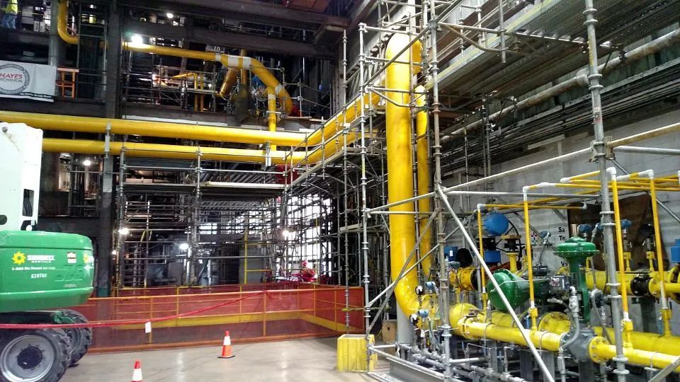 Industrial plant interior with yellow pipes, machinery, and safety barriers around equipment.