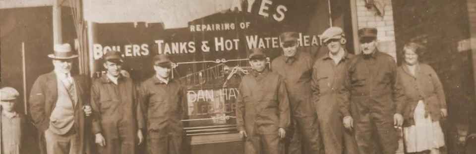 Group of men posing outside a boilers, tanks, and hot water heater shop sign, early 1900s.