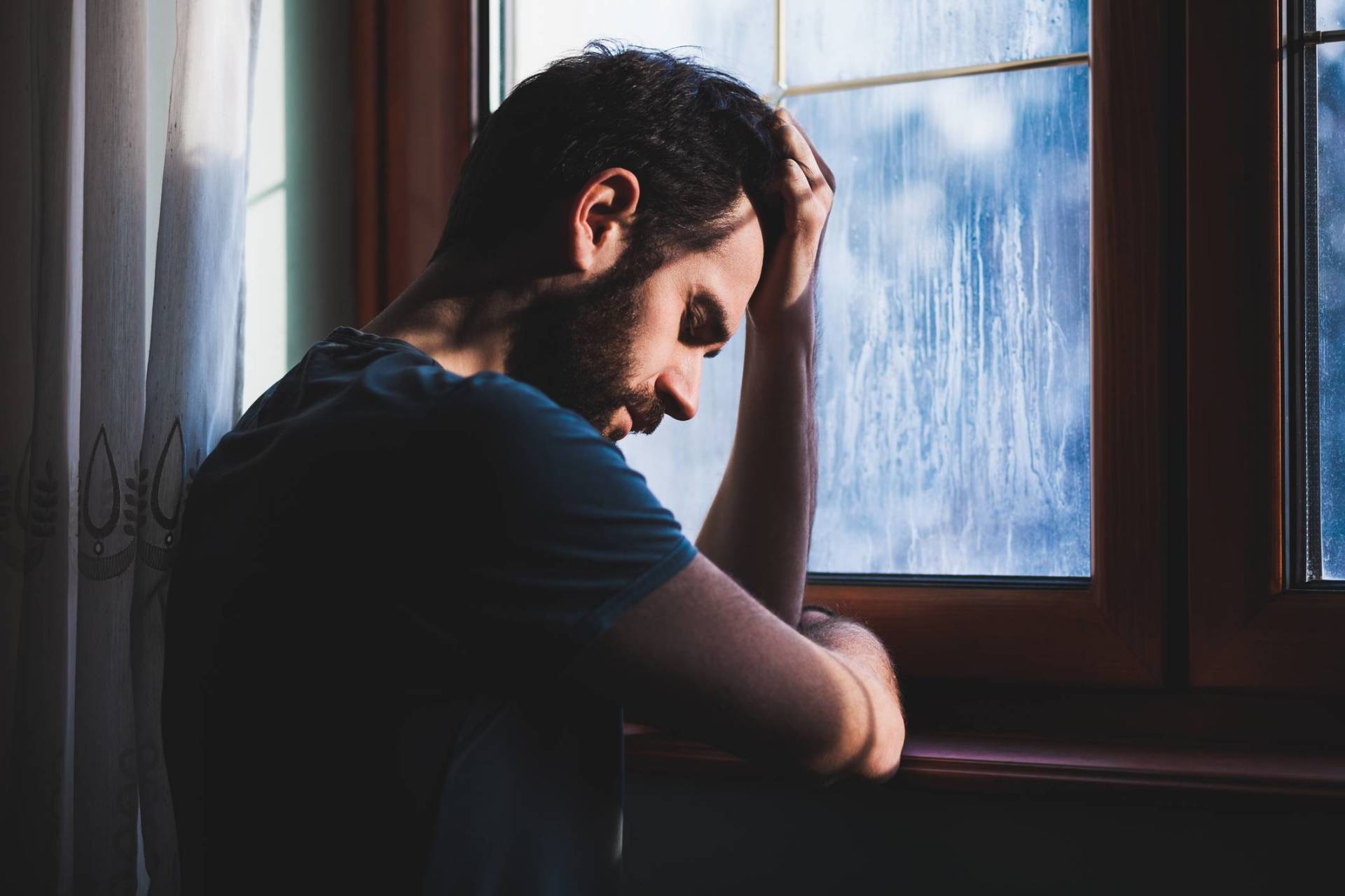 A man holding his head and looking sad leaning on a windowsill while it rains outside