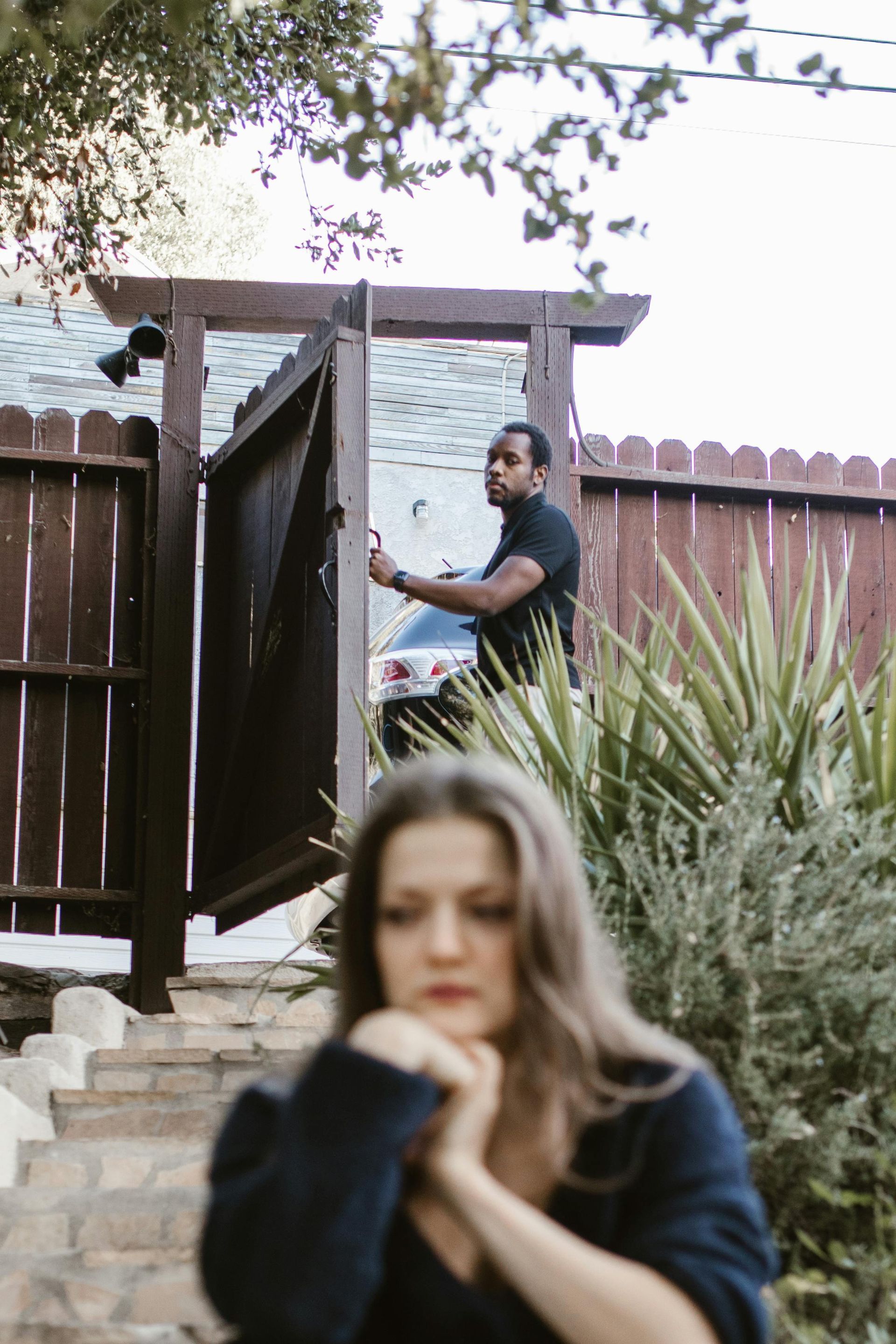Woman at bottom of stairs, man by open gate. Wooden fence.