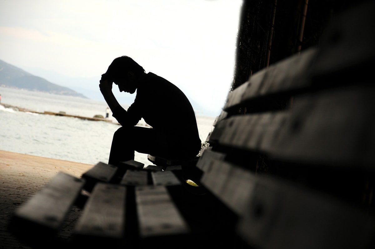 A man sitting on a bench in the shadow and holding his head