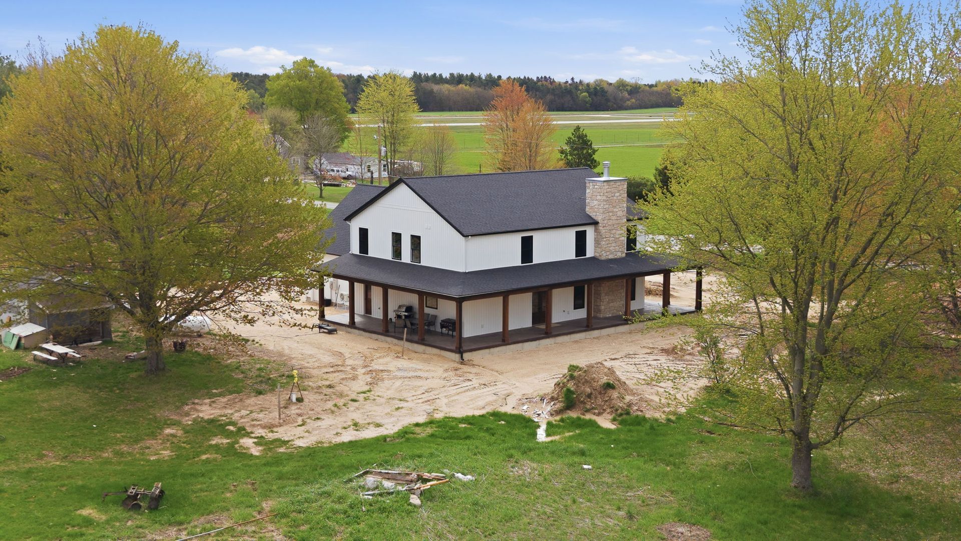 White two-story house with a black roof on a rural lot, surrounded by trees and open fields