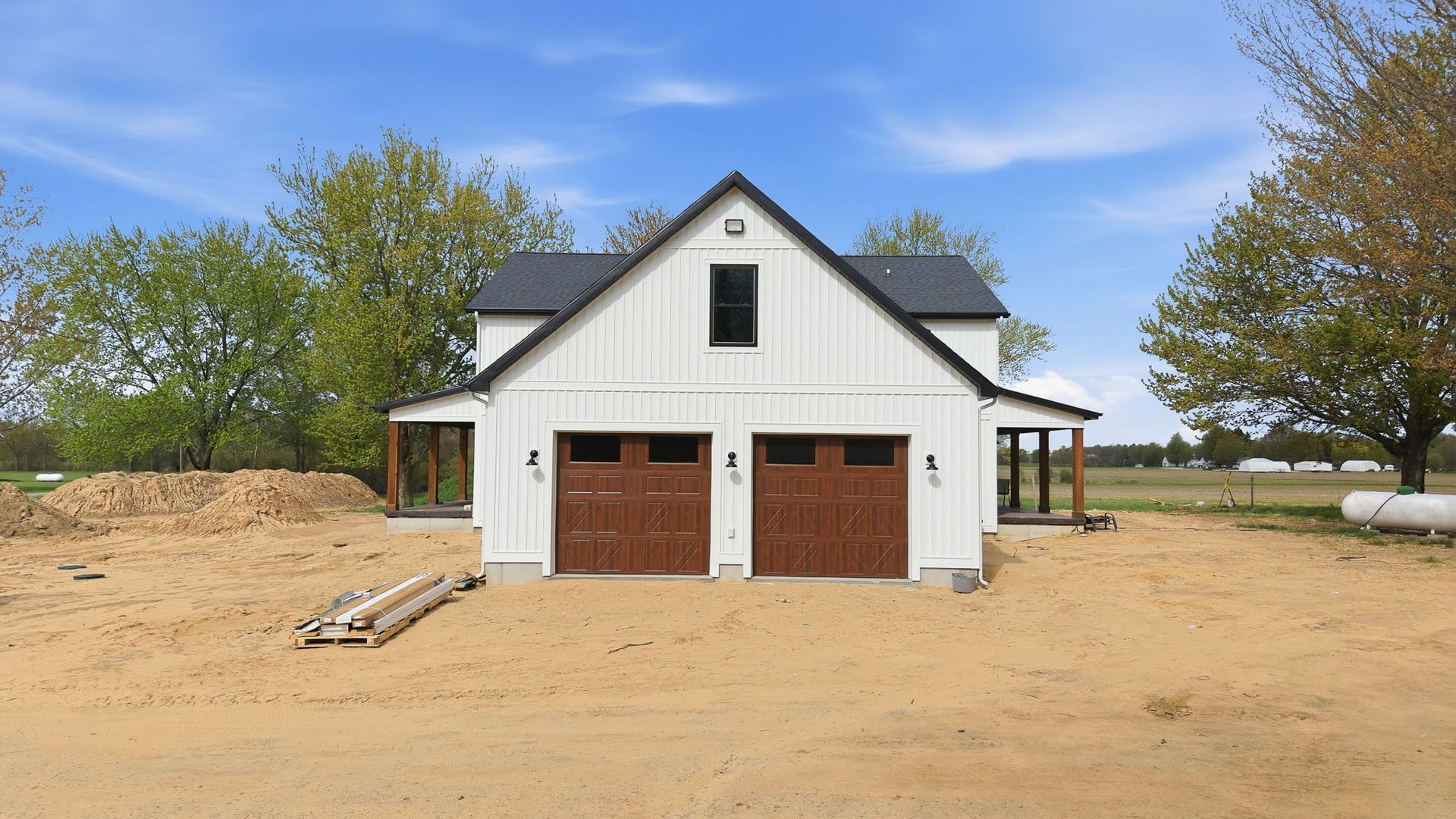 White barn-style house with brown garage doors on a sandy lot under a blue sky