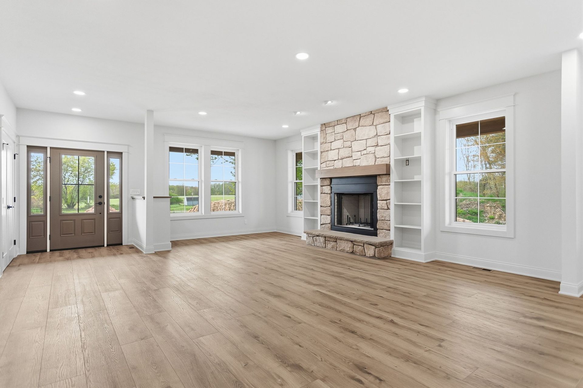 Bright empty living room with hardwood floors, stone fireplace, and large windows.