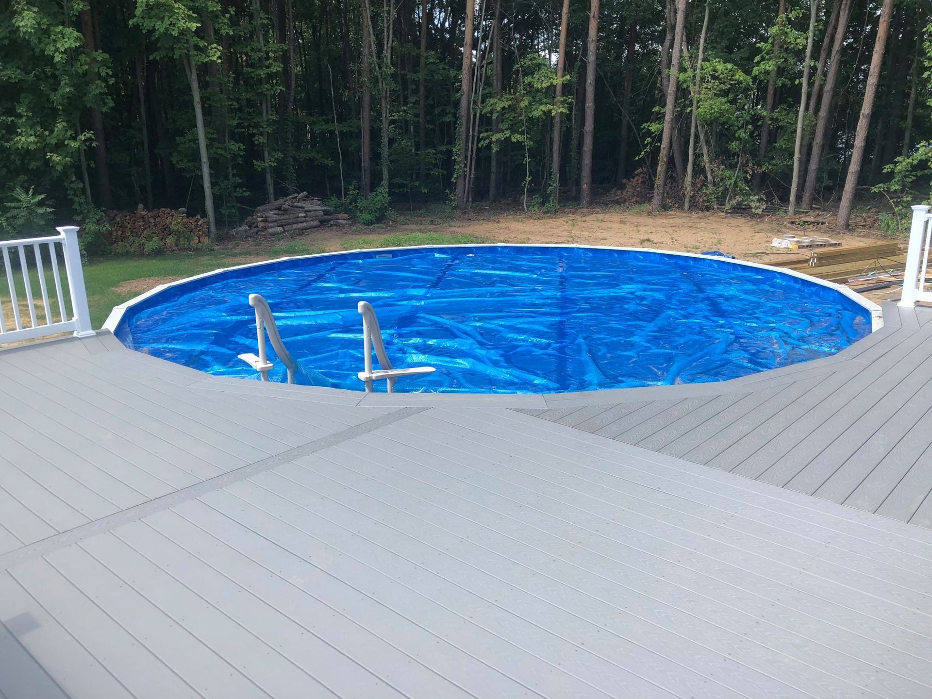 A blue, circular above-ground pool sits in a backyard, surrounded by a gray composite deck and a line of tall trees.