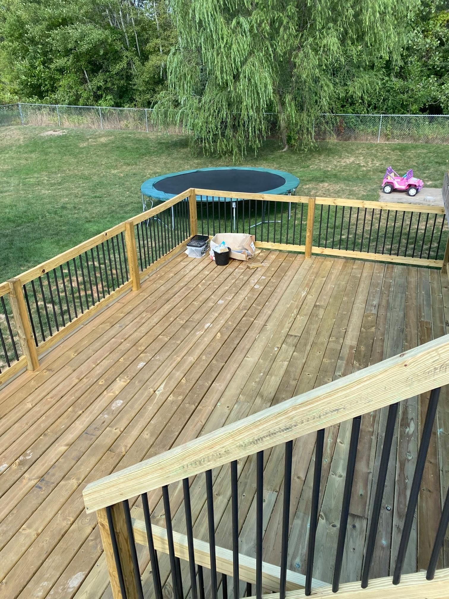 A wooden backyard deck with railings overlooks a grassy lawn containing a small trampoline and a pink toy car.