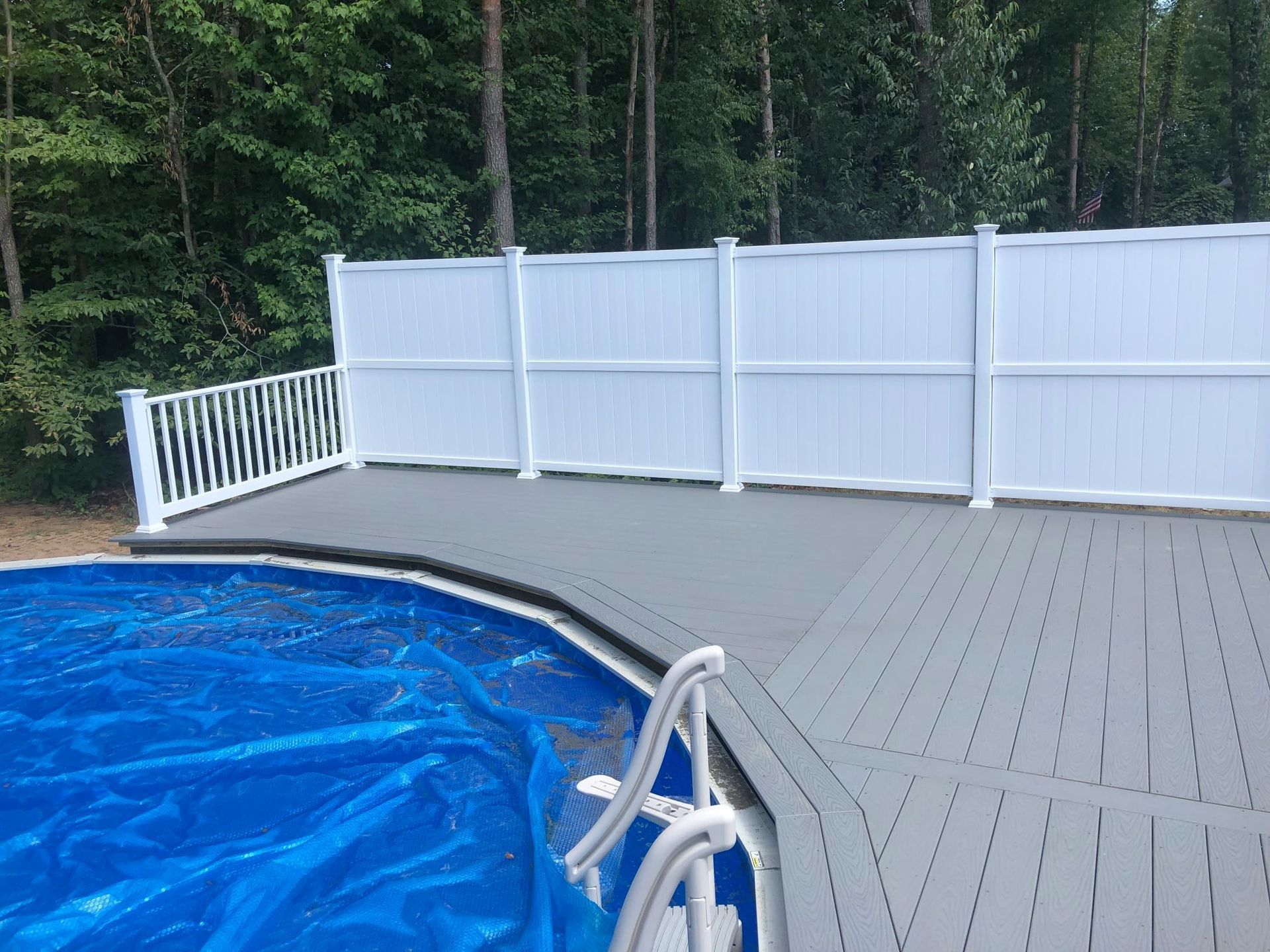 A grey composite deck next to an above-ground pool covered with a blue solar blanket, set against a white vinyl fence.
