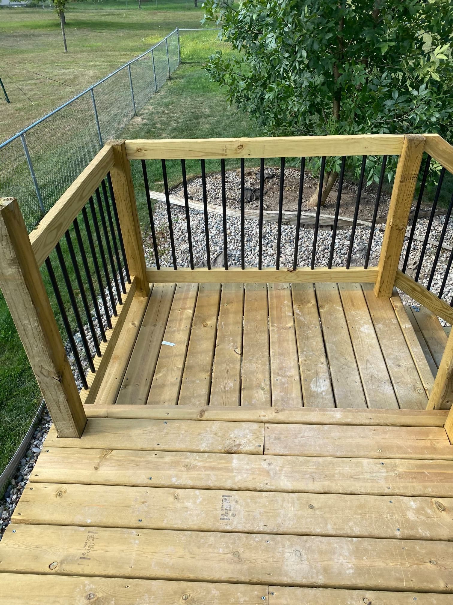 A wooden deck landing with black metal railing, overlooking a gravel yard with a chain-link fence and tree.
