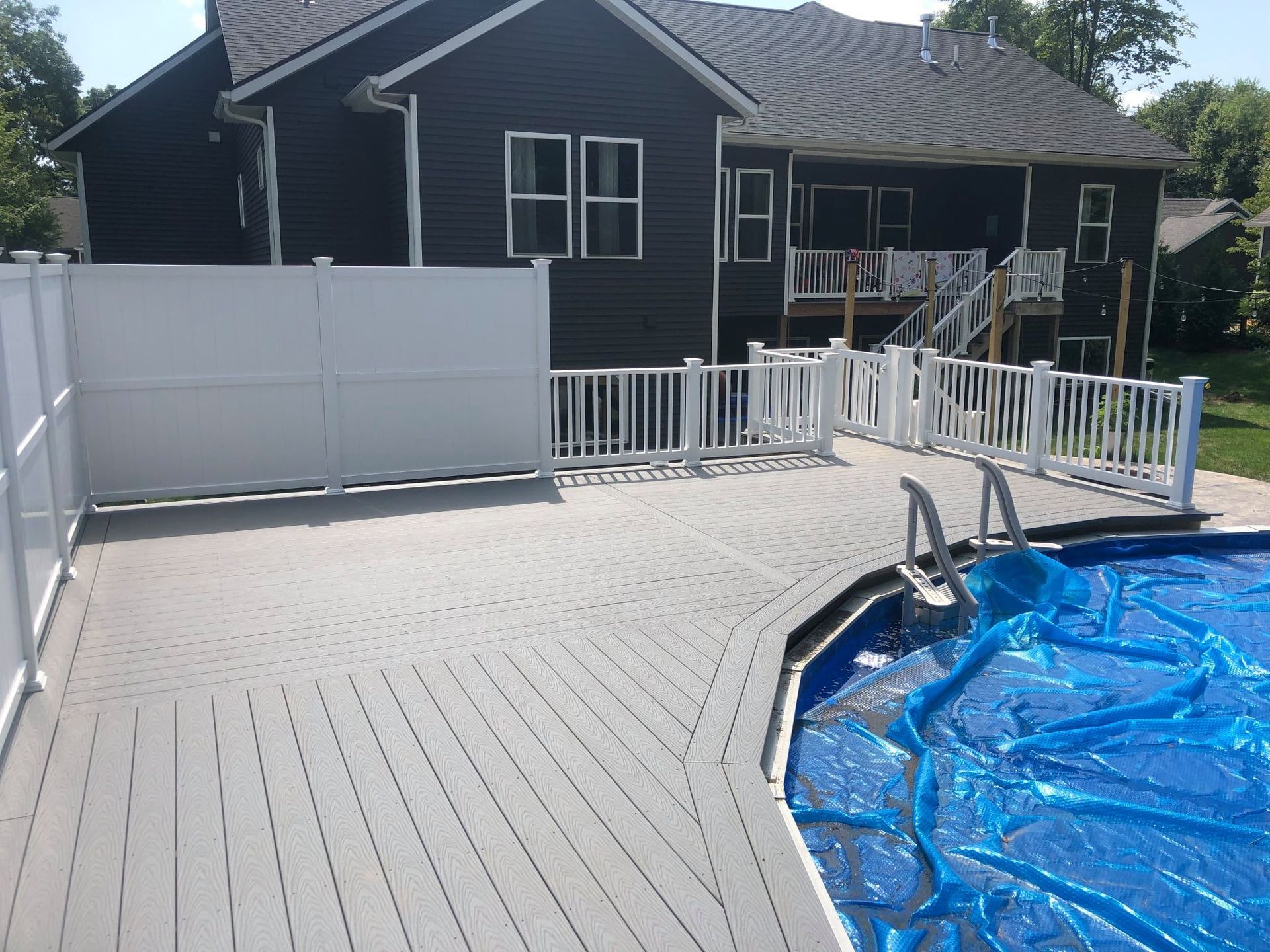 A gray composite deck with white railings and privacy fencing surrounds a backyard swimming pool with a blue cover.