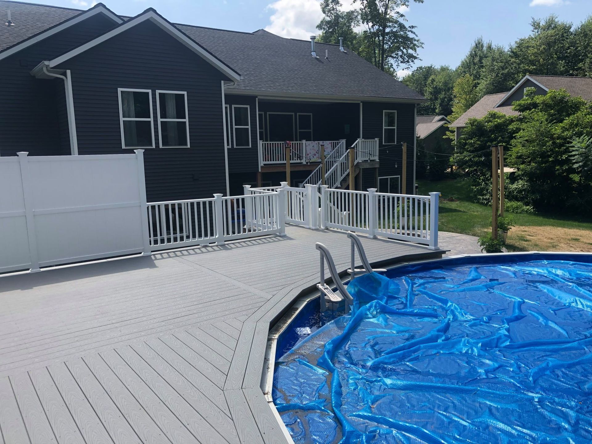 A gray composite deck surrounding an above-ground pool covered with a blue solar blanket near a dark-sided house.