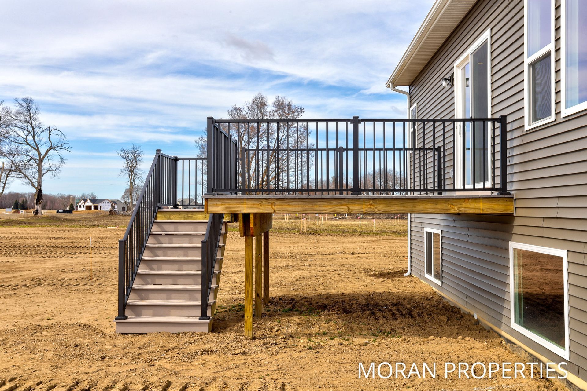 A ground-level view of a new wooden deck with black railings and stairs attached to the side of a gray-sided house.
