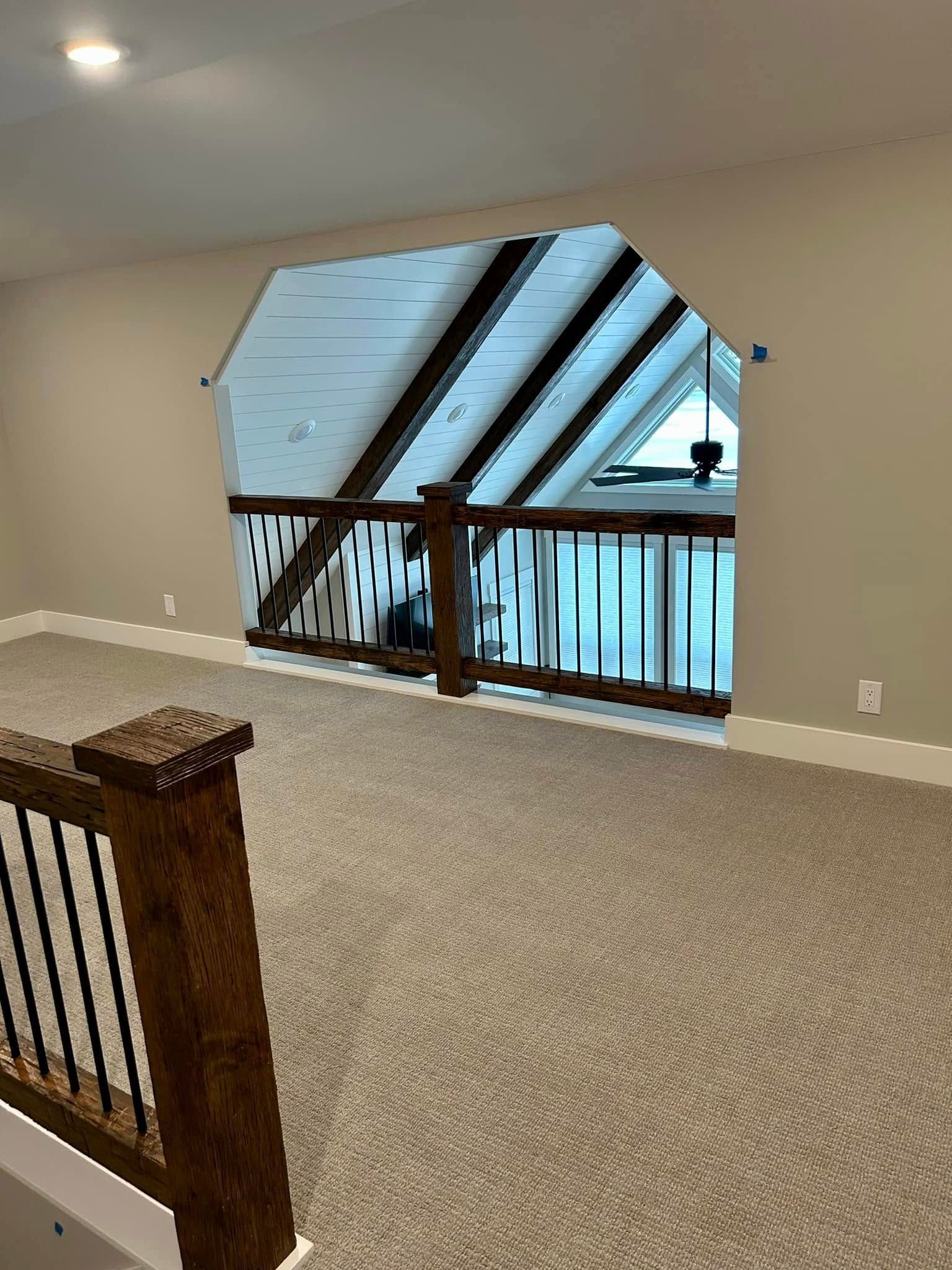 A carpeted loft space with wooden railings overlooking an opening with exposed beams and a triangular window.
