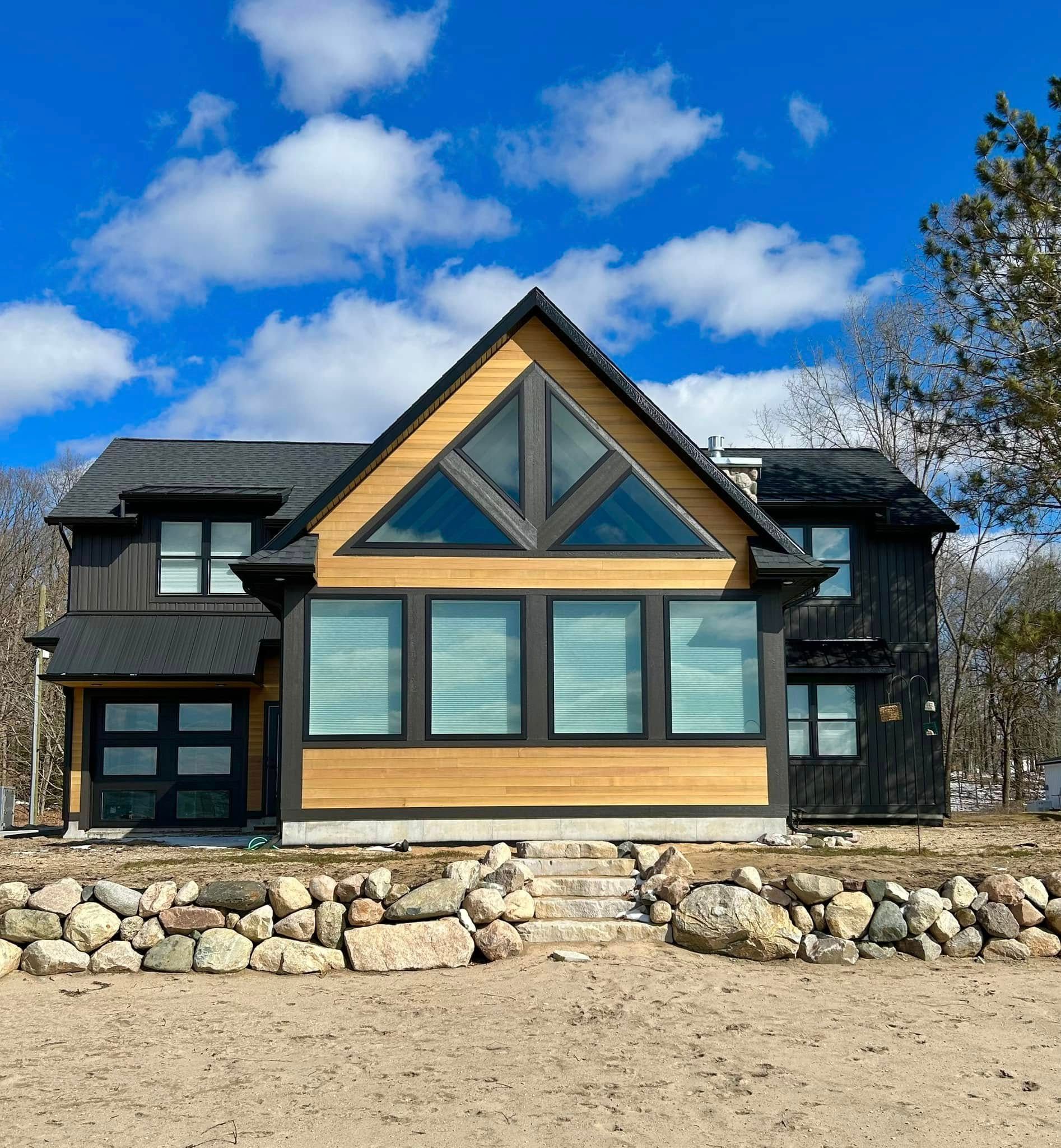 A modern A-frame house with light wood siding and dark trim sits on a sandy beach behind a stone retaining wall.