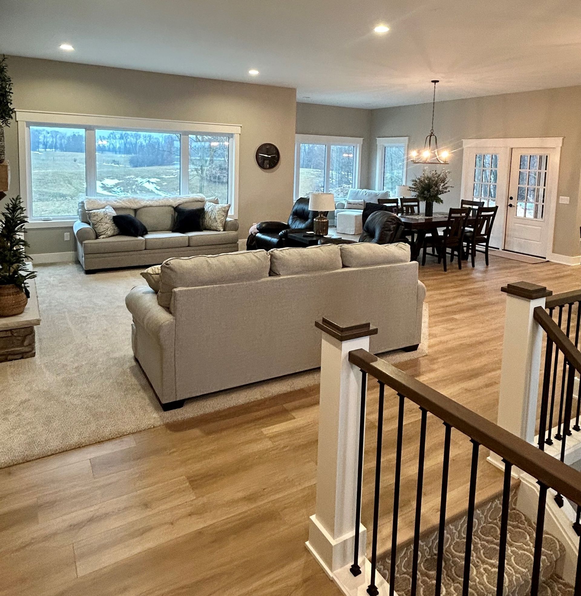 A bright, open-concept living and dining room with neutral furniture, hardwood floors, and a scenic view through windows.