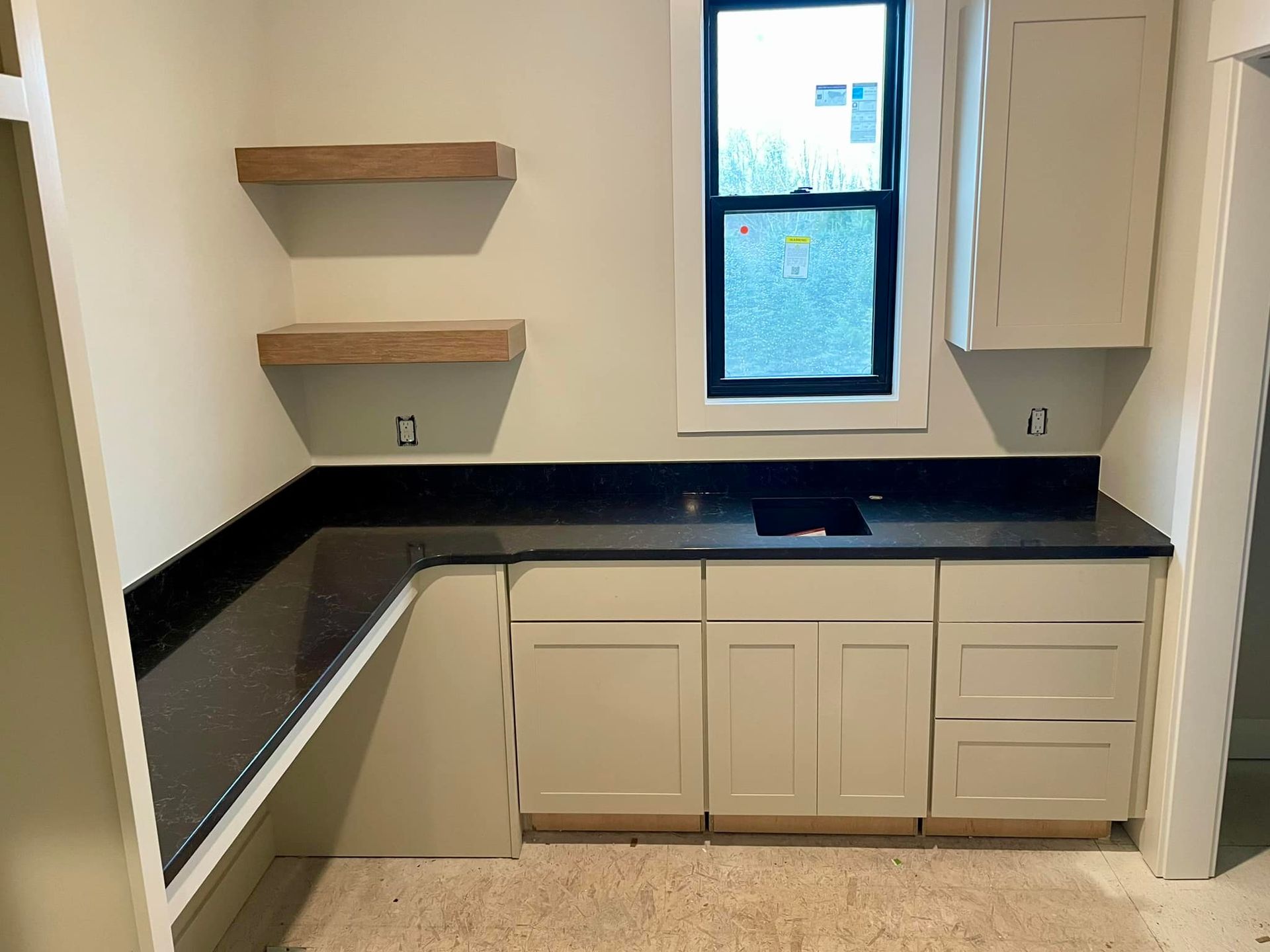A kitchen corner with off-white cabinets, a black countertop, a sink, a window, and two floating wooden shelves.