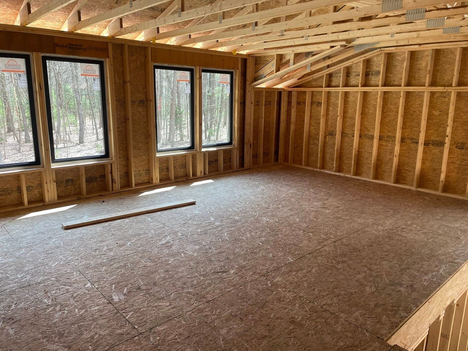 Interior of a room under construction with wood-framed walls, plywood flooring, ceiling trusses, and three windows.