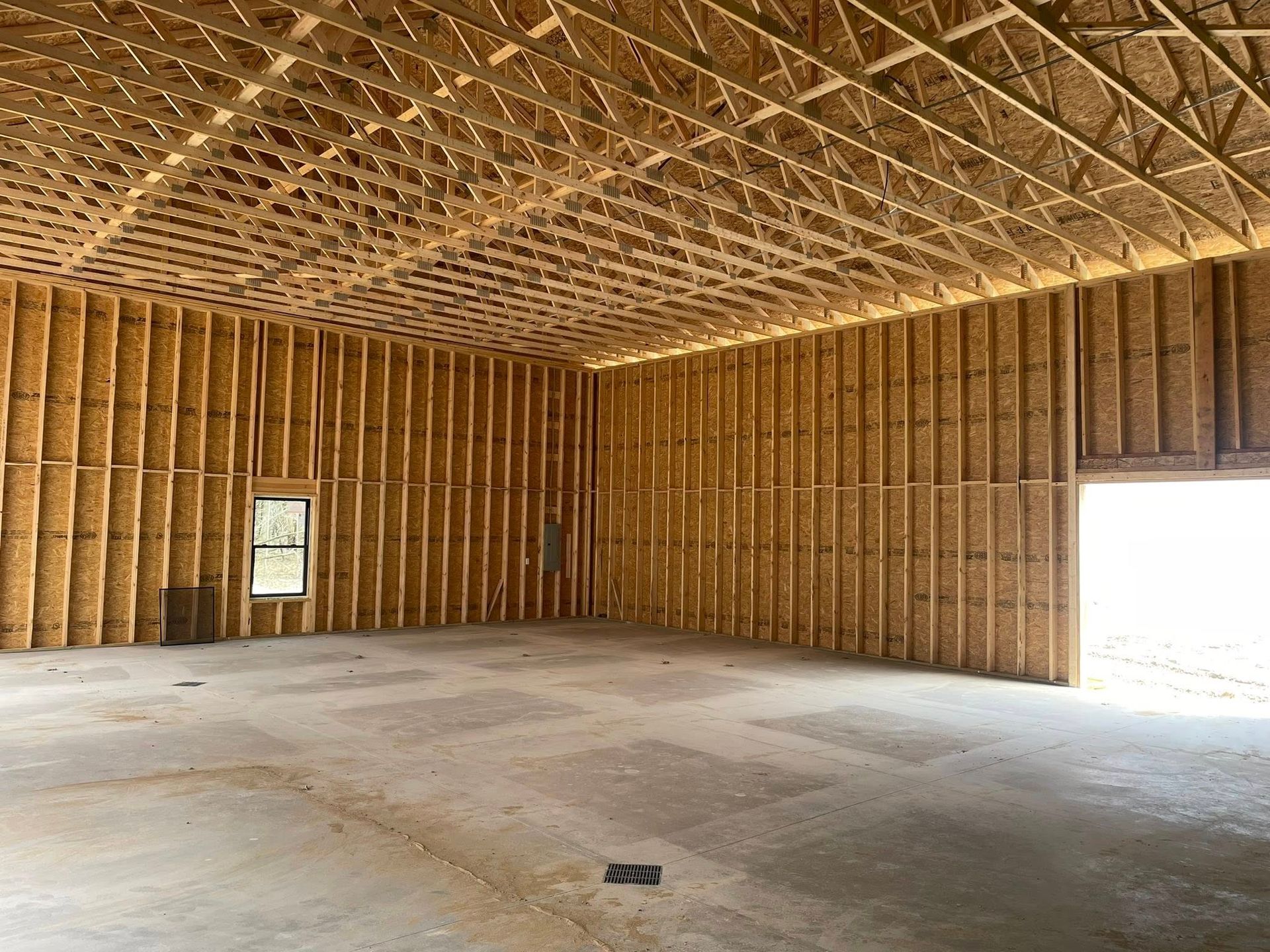 The interior of a large, unfinished building with exposed wood wall studs, a wooden truss ceiling, and a concrete floor.