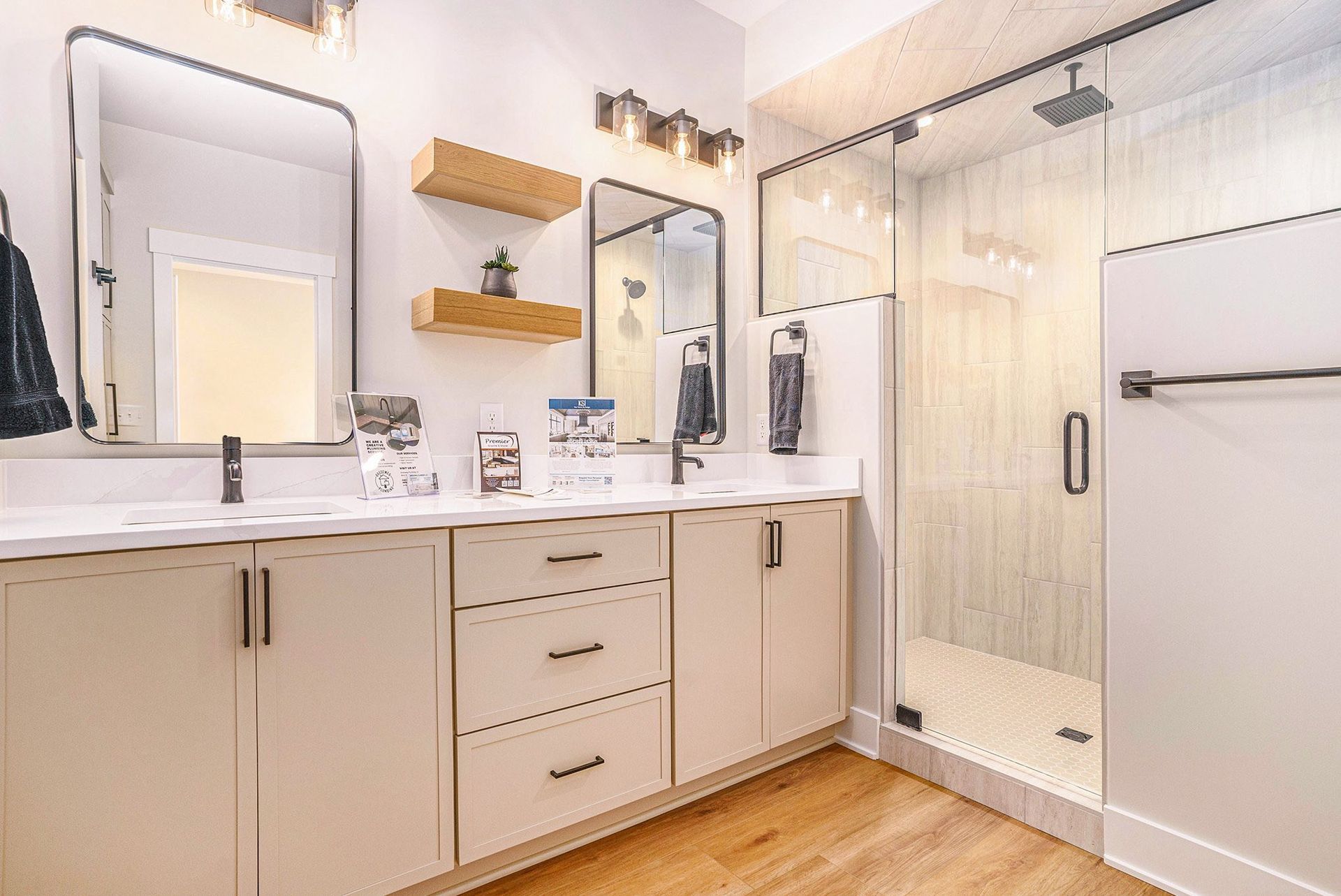 Modern bathroom with double vanity, two mirrors, wood shelves, and a walk-in glass shower with light-colored tile.