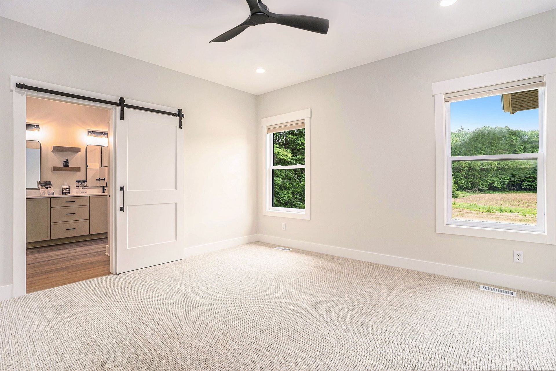 A bright bedroom with beige walls, patterned carpet, a ceiling fan, a barn door to a bathroom, and two large windows.