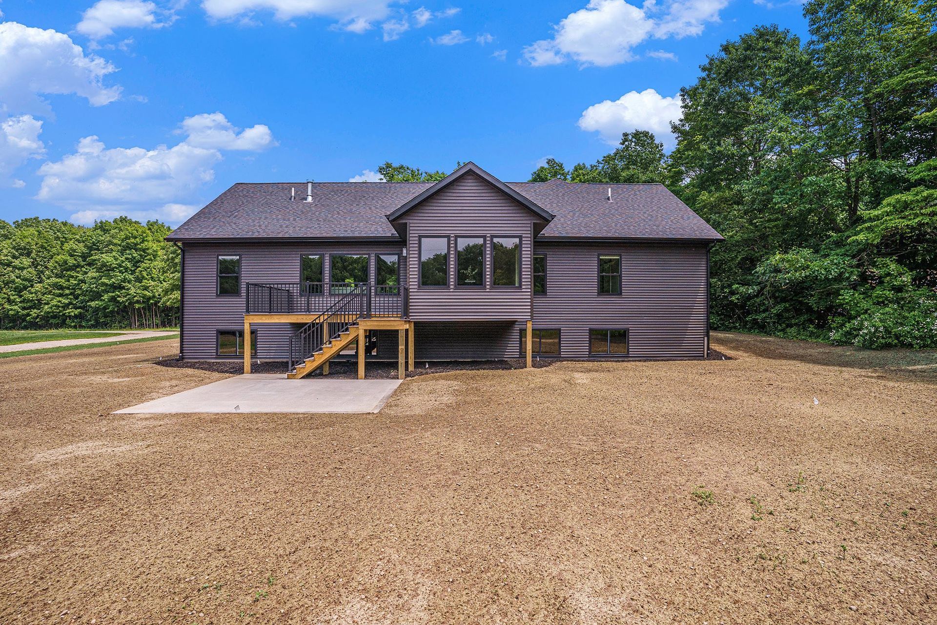A dark-sided house with a back deck, stairs, and a concrete patio set in a rural backyard with trees under a blue sky.