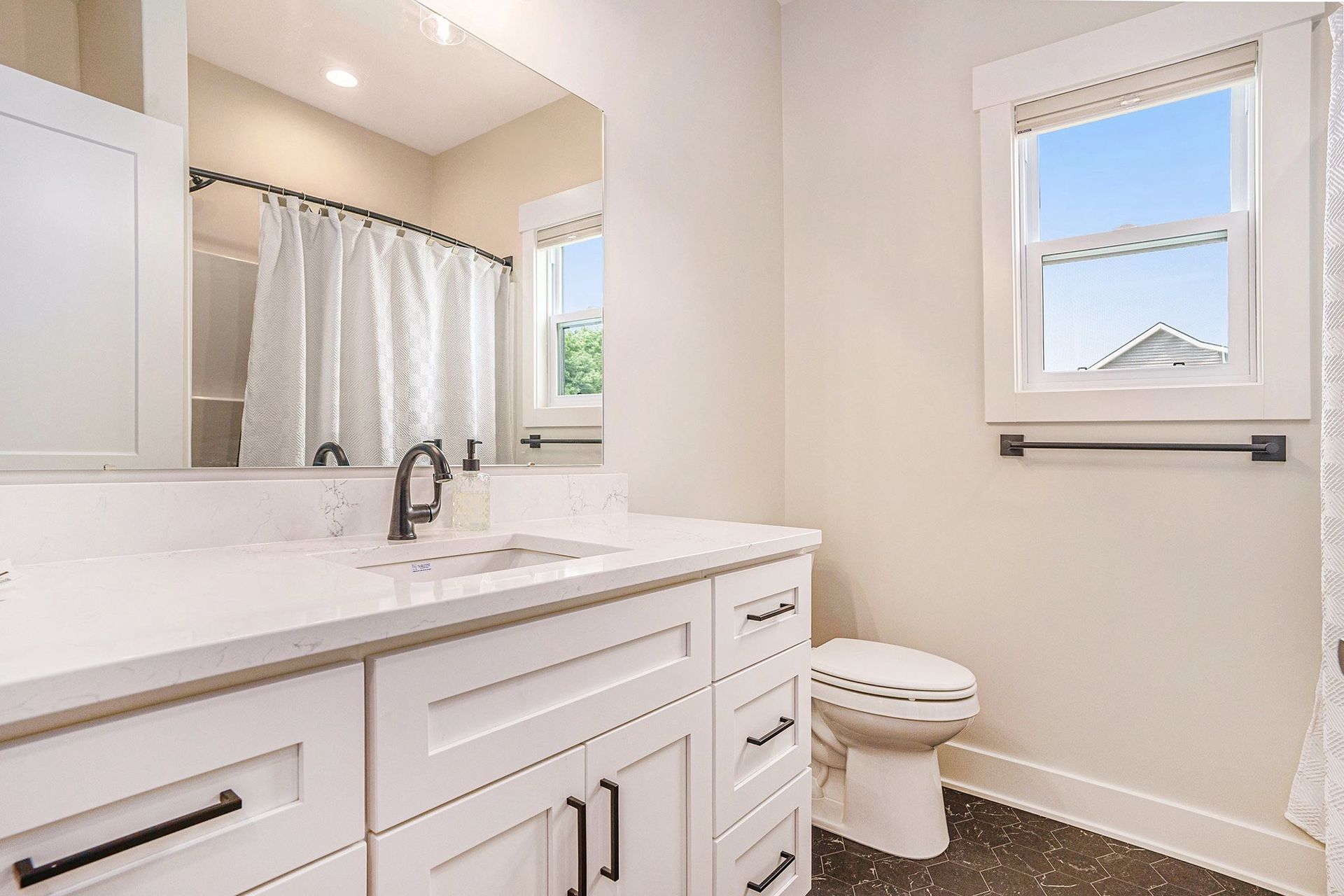 A bright, modern bathroom featuring a white vanity with black hardware, a shower with a curtain, and a window above a toilet.
