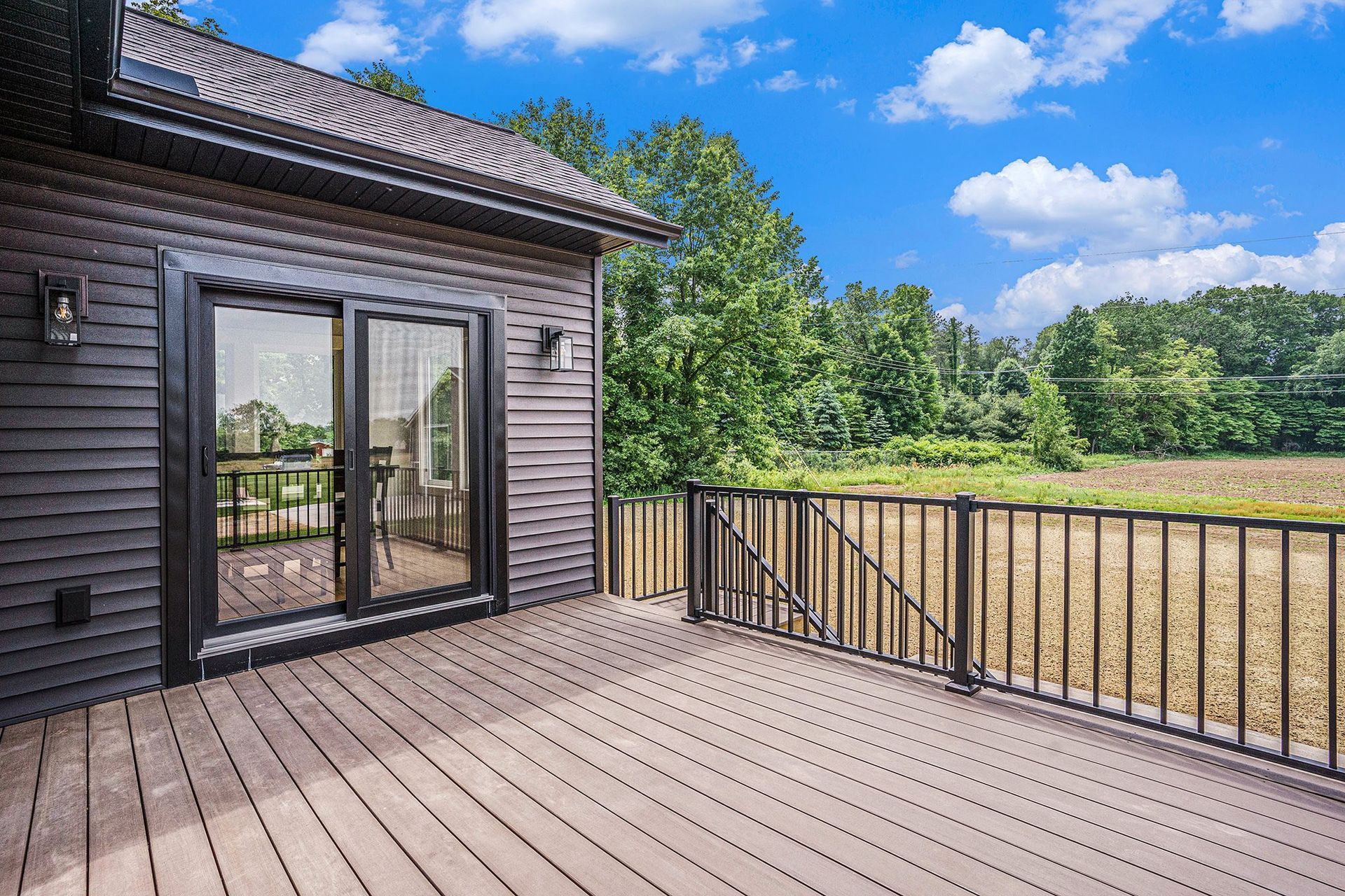 Dark wood-sided house with a sliding glass door and deck overlooking a sunny, grassy field under a bright blue sky.