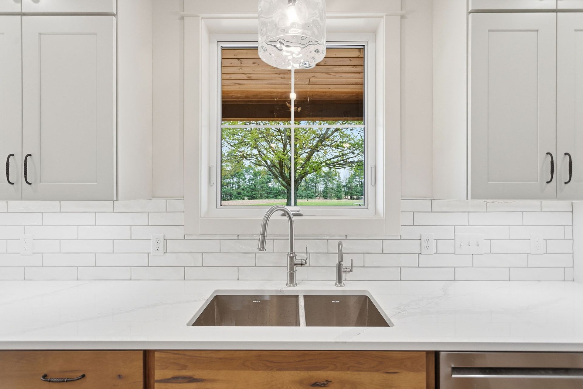Bright white kitchen with centered window above a double sink and chrome faucet, wood blinds, and subway tile backsplash