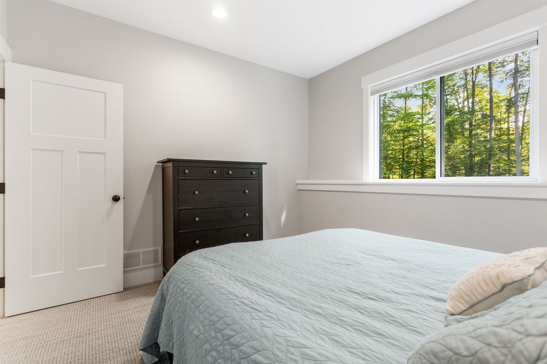 A bedroom featuring a blue quilted bed, a dark wood dresser, a white door, and a window with a view of green trees.