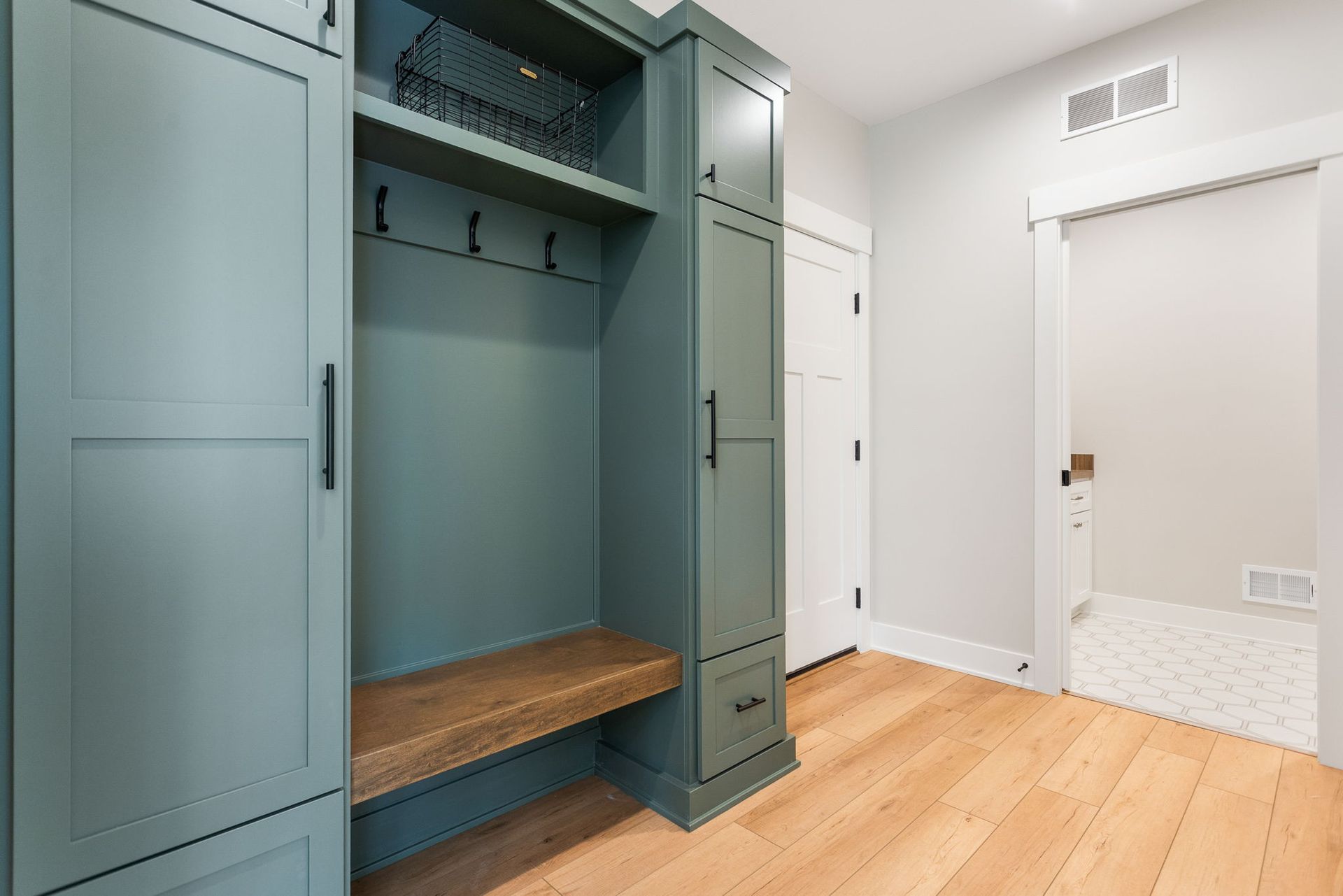 A mudroom with muted green cabinets, a wood bench, coat hooks, and an open doorway leading to a tiled room.