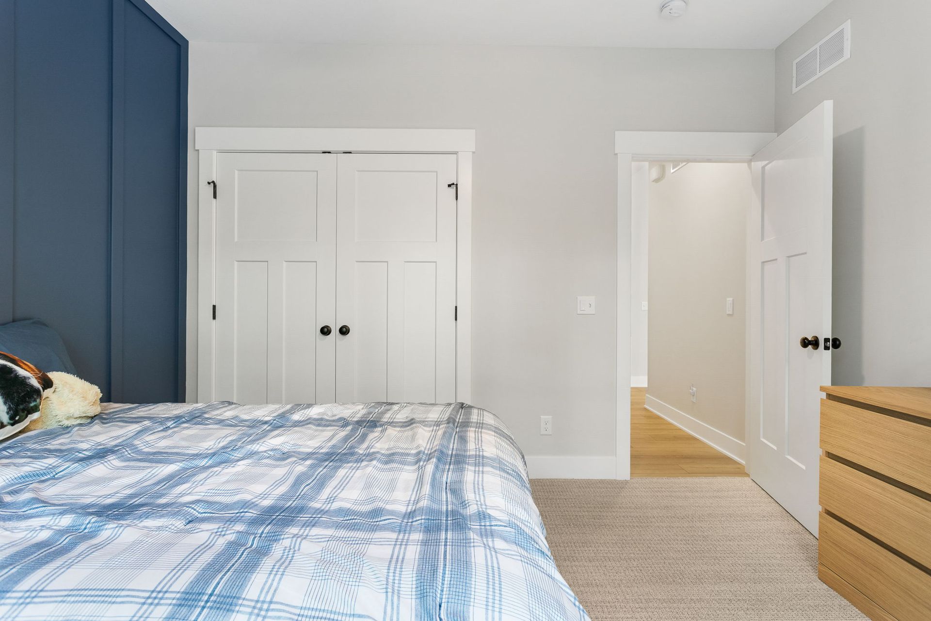 A bedroom with a blue accent wall, a plaid bedspread, white double closet doors, an open doorway, and a wooden dresser.
