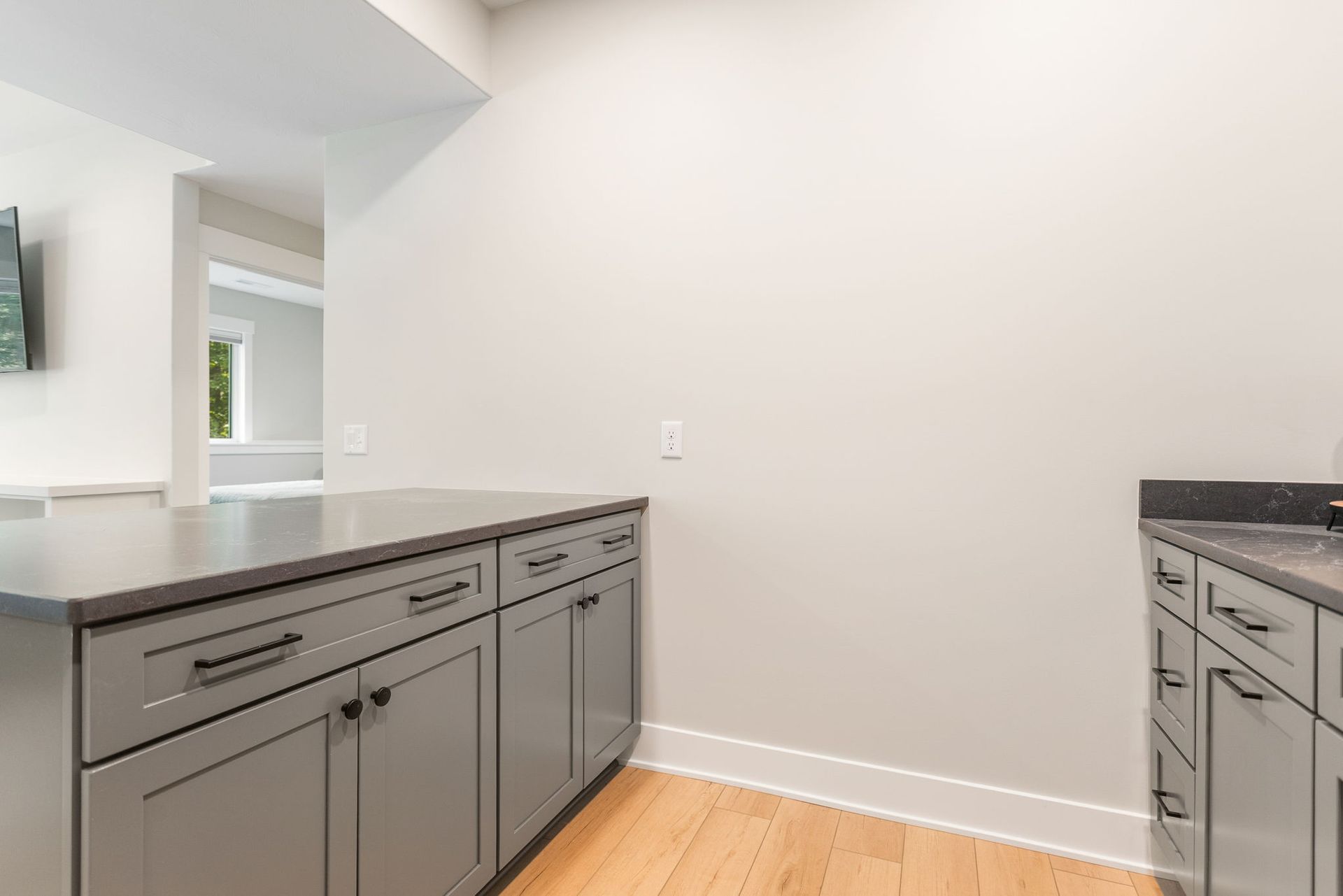 Gray kitchen cabinets with a dark countertop and light wood flooring in a modern, neutral-toned room.