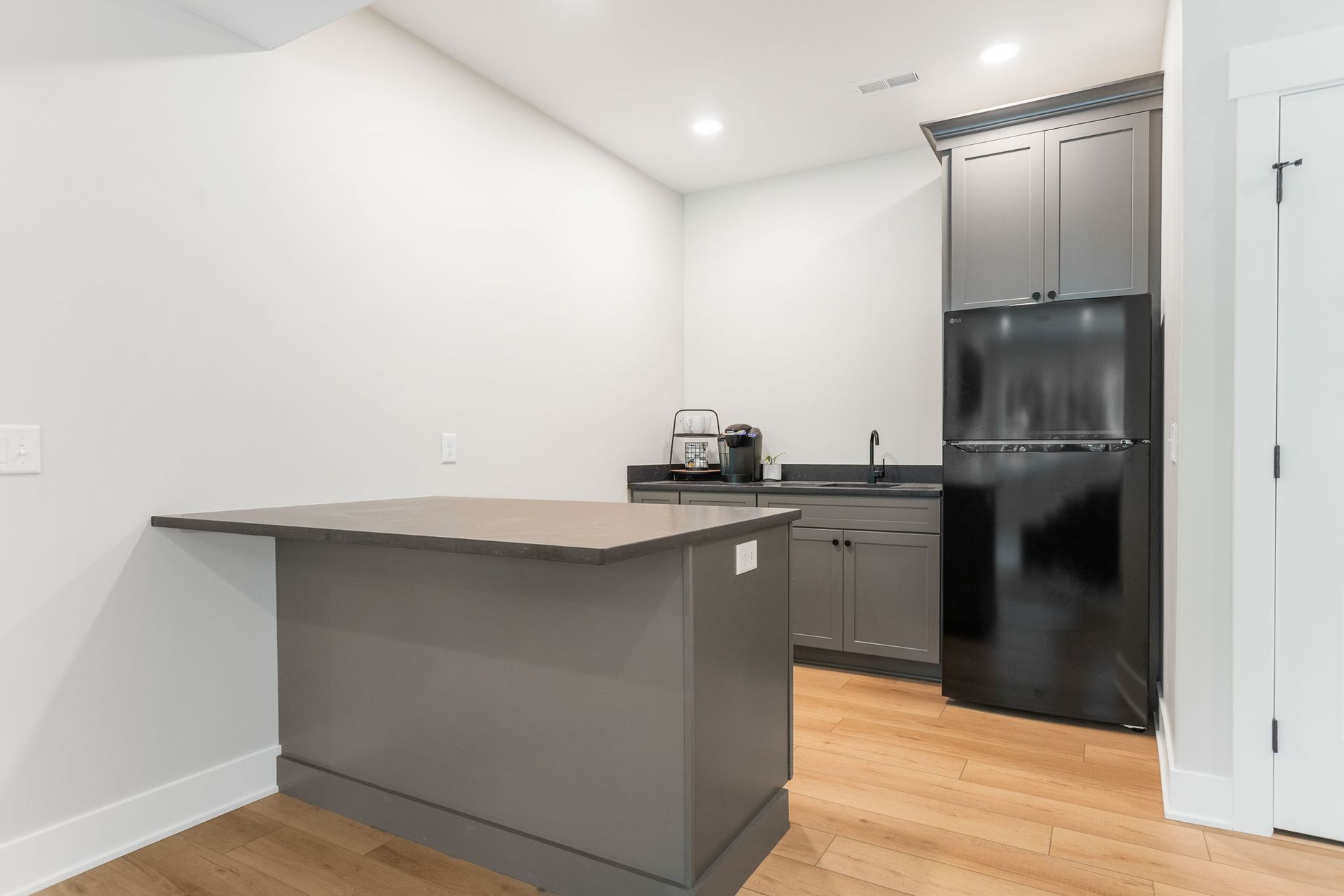 A small kitchenette featuring dark grey cabinets, a matching kitchen island with a countertop, and a black refrigerator.