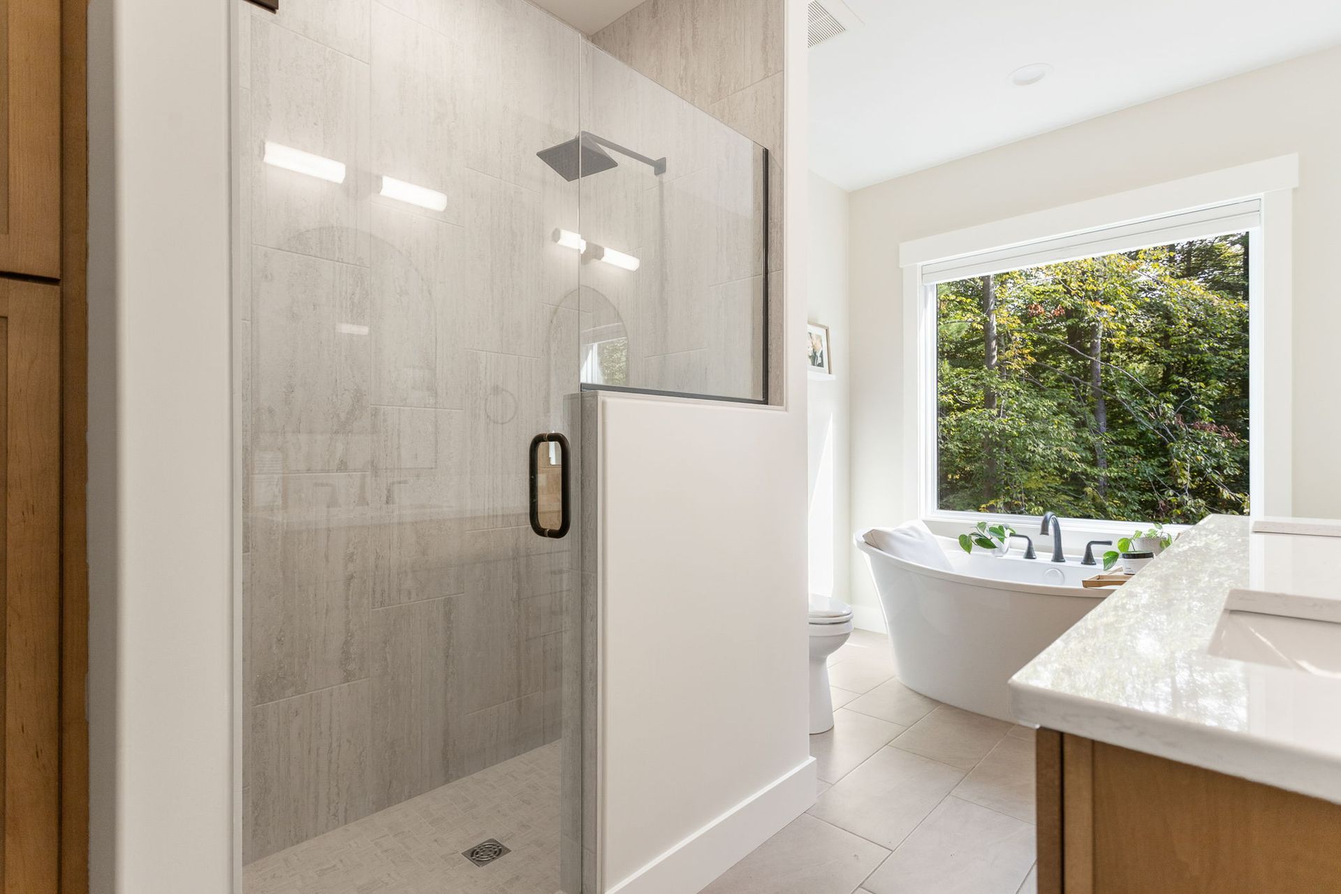A modern bathroom with a glass-enclosed shower, a freestanding tub by a window, and wood-toned cabinetry.