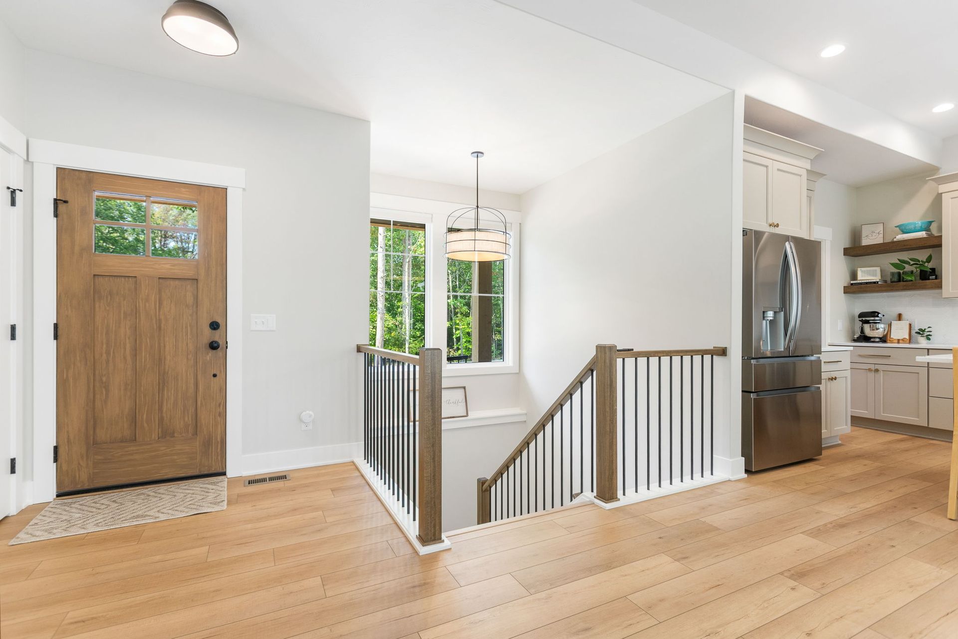 A modern entryway featuring a wood front door, light hardwood floors, a staircase with metal railings, and a kitchen view.