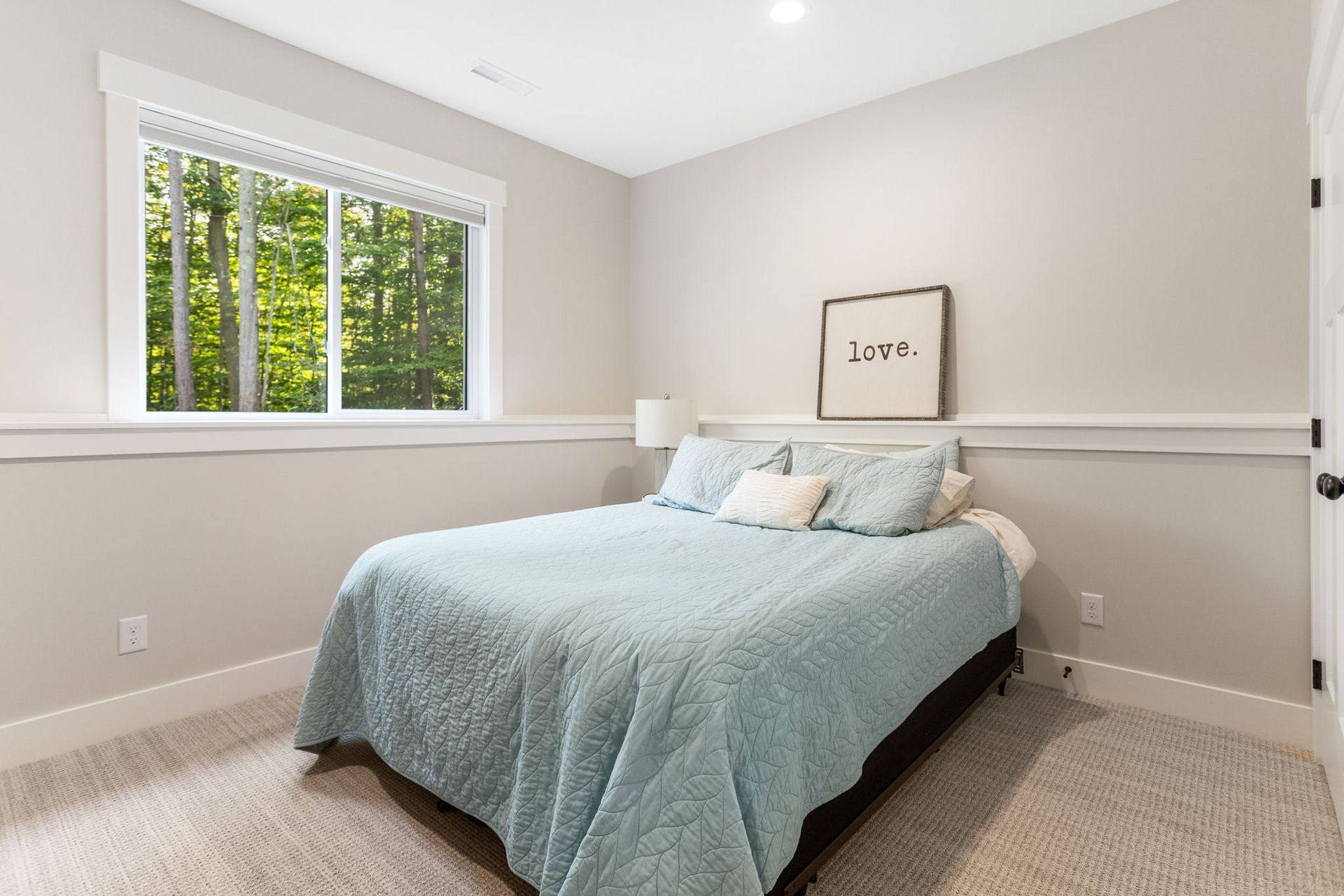 A tidy bedroom with a light blue bedspread, a window overlooking trees, beige walls with wainscoting, and carpeted floors.