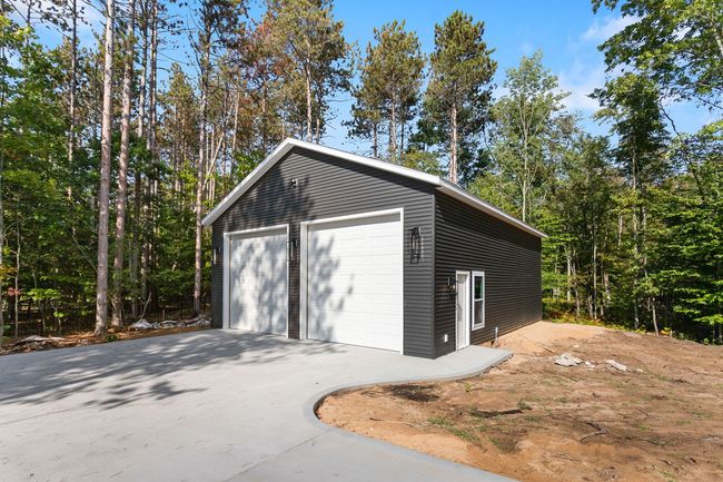 A modern dark-gray detached garage with two white doors and a concrete driveway, surrounded by a forest.